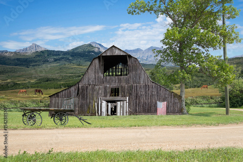 horizontal image of an old run down barn with an American flag hanging from it and a wooden hauling wagon sitting in the yard with beautiful mountains in the background with horses grazing in pasture.