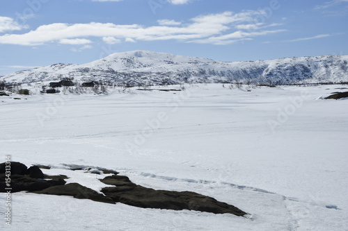 View over a frozen lake with snowmobile tracks and a Sami village and a snowy mountain