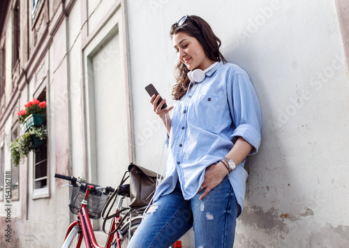 Portrait of beautiful young woman with bicycle in old town leaning on wall and looking at mobile phone.