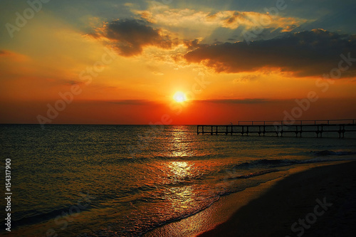 Golden natural sea sunset view of small bridge at horizon and red sky landscape. Sunset reflection in nature with sun in clouds above black sea scenery.  