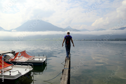 Travel to Sankt-Wolfgang, Austria. The young man are walking on a bridge with the view on the lake near to mountains in the cloudy weather.