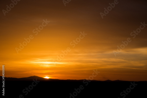 Beautiful sunset with half of the sun coming down behind some mountains and orange tones in the sky and clouds © Massimo