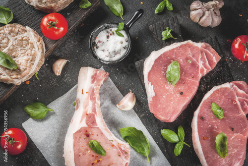 Raw organic meat. Selection of several types of red meat: pork cutlet on bone, pork steaks and chicken cutlets in grid of pork fat. With ingredients for cooking, gray stone table top view