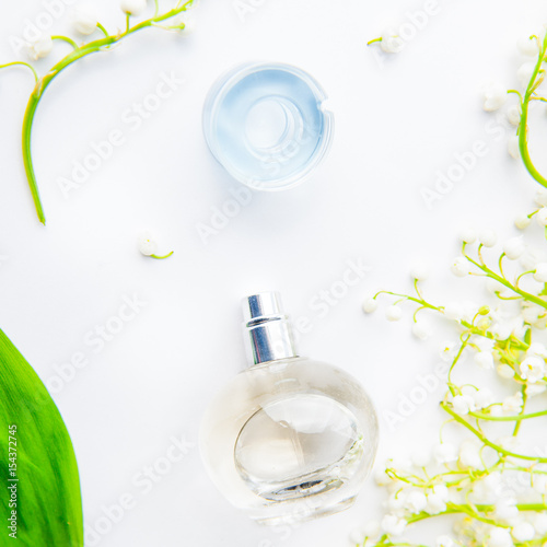 Square Beauty concept card. Flat lay with Orbicular perfume bottle surrounded by fresh lilies of the valley, may-lily flowers and green leaf on the white background. Top view. Selective focus