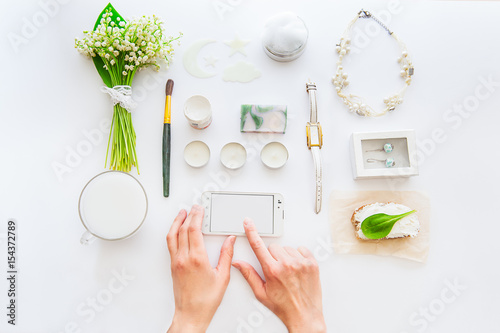 Beauty blog concept. Female hands touch smartphone screen on the background of styled greenery and white datails and accessories on white workplace. Flat lay, top view fashion trends. Space for text photo