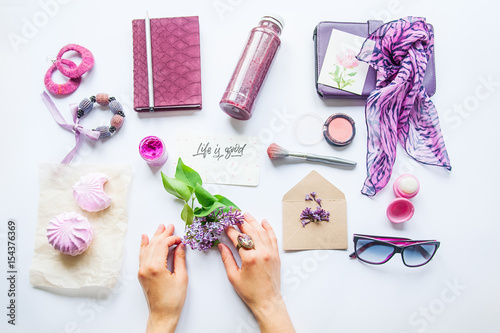 Beauty blog concept. Lilac colour. Female hands keep lilac flower among styled accessories: sunglasses, bijouterie, cosmetics and sweets on white background. Flat lay, fashion background. photo