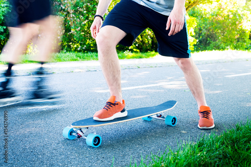 Young man riding on a longboard. Longboard on the road in sunny weather. People around skateboard. Pathway in a park. Urban scene.