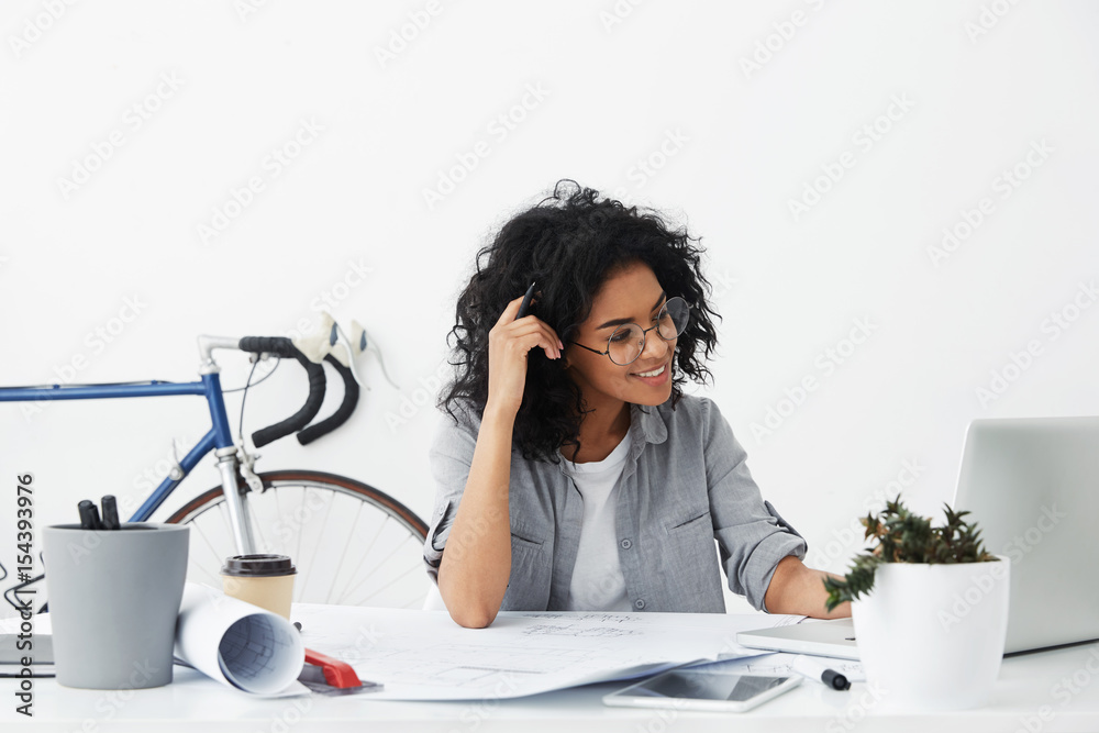 Φωτογραφία Positive young beautiful american woman wearing stylish shirt and big round glasses making drawings and studying sketches working with modern gadgets in office isolated over white background
