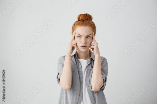 Fototapeta Studio shot of serious redhead young Caucasian woman holding fingers on her temp