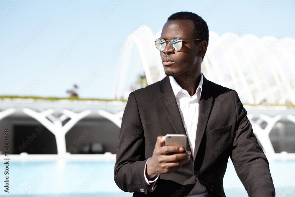 Confident young black businessman formally dressed having round stylish shades holding smart phone messaging online looking aside isolated over urban background. Modern technologies and businesspeople Confident young black businessman formally dressed having round stylish shades holding smart phone messaging online looking aside isolated over urban background. Modern technologies and businesspeople