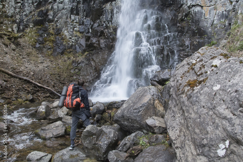 Obraz premium A backpacker with a backpack and a rug near the big waterfall among the rocks