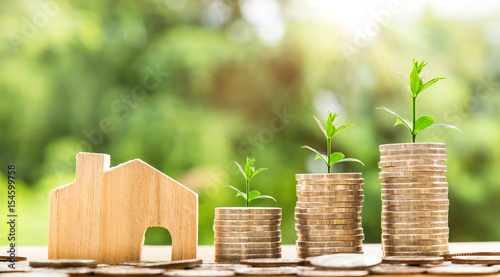 step of coins stacks with tree growing on top and wooden house model on table, nature background, money, saving and investment or family planning concept, over sun flare silhouette tone.