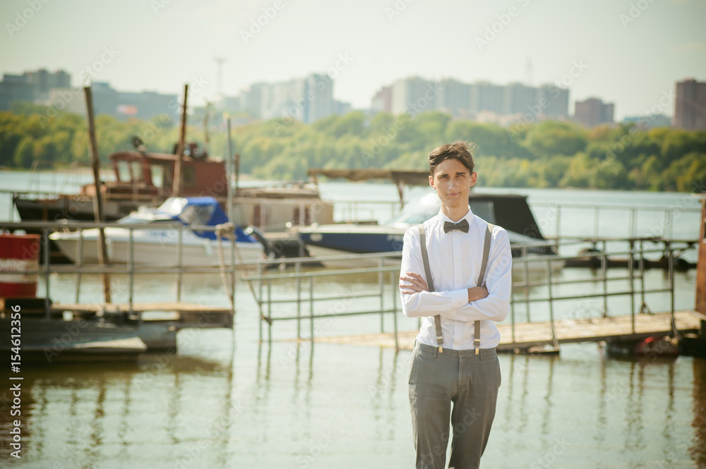 young skinny man, elegantly stylishly dressed in white shirt, gray trousers with suspenders and bow tie. Portrait young guy on pier on background of river and boats
