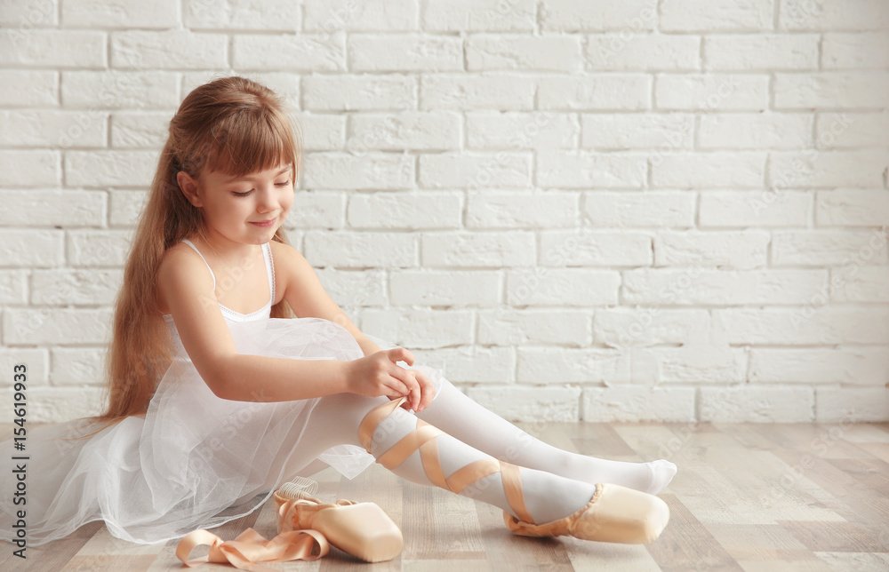 Naklejka premium Beautiful little ballet dancer tying pointe shoes in light dance studio
