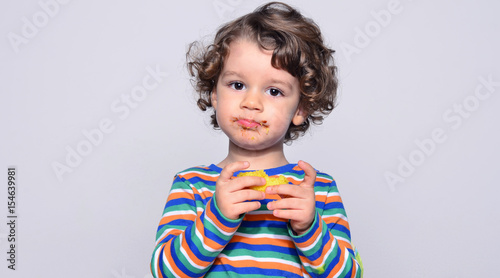 Kid getting messy while eating a chocolate cake. Beautiful curly hair boy eating sweets. Toddler in high chair being hungry stuffing his mouth with cake 
