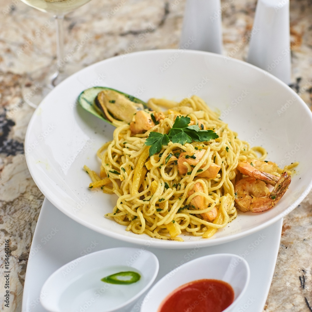 Flat egg noodles with seafood served in big white ceramic round plate with glass of white wine on marble table