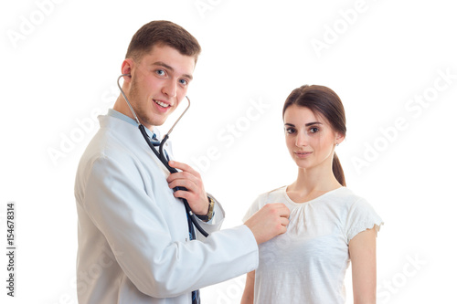 young smiling doctor looks straight stands near girls and listens to her heart with a stethoscope
