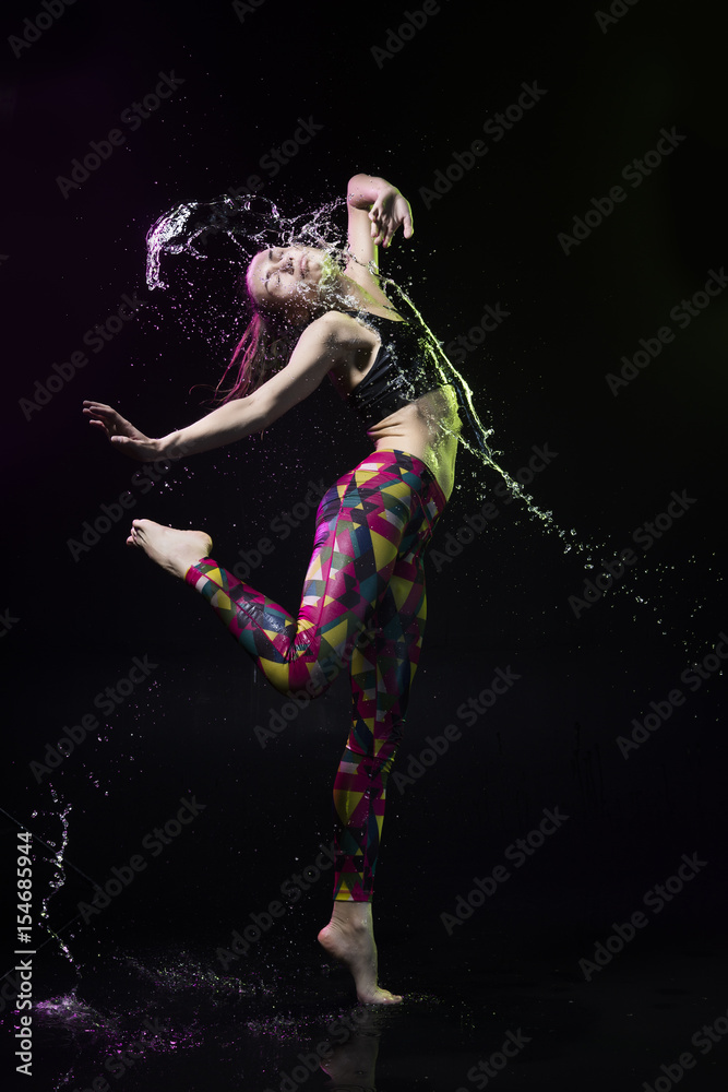 Obraz premium The girl dances on the floor covered with water on a black background and water splashes around her