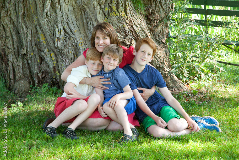 Fototapeta premium Happy mother sat in a park with her arms around her three young sons.