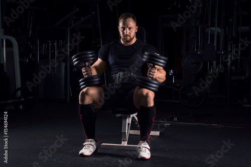 Impressive athlete sits on a bench with two dumbbells on his legs