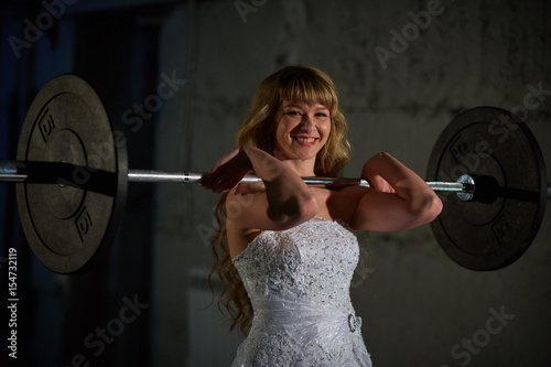 Beautiful and muscular bride blonde in a wedding dress doing an exercise with a barbell in a gym, crossfit photo