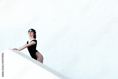 A beautiful young free sexy girl dressed in bodysuit in pantyhose and sunglasses stands in shoes, with a smart ass and long legs against a white wall background