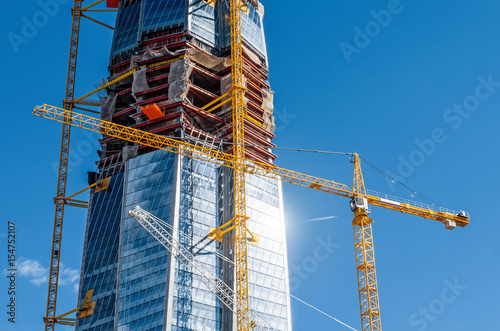 Scale active skyscraper building, glare from the sun on the windows, cranes against the blue sky.