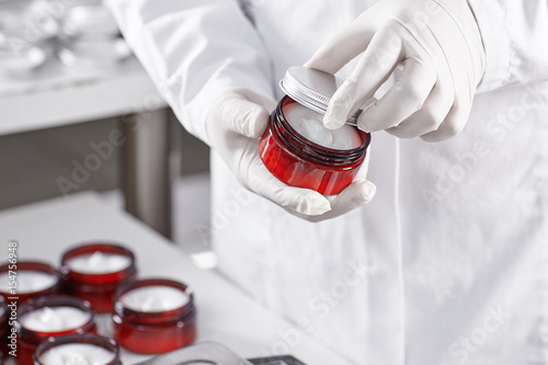 Foto Factory worker in white gown and gloves holding red container with skin care product touching it with finger testing quality