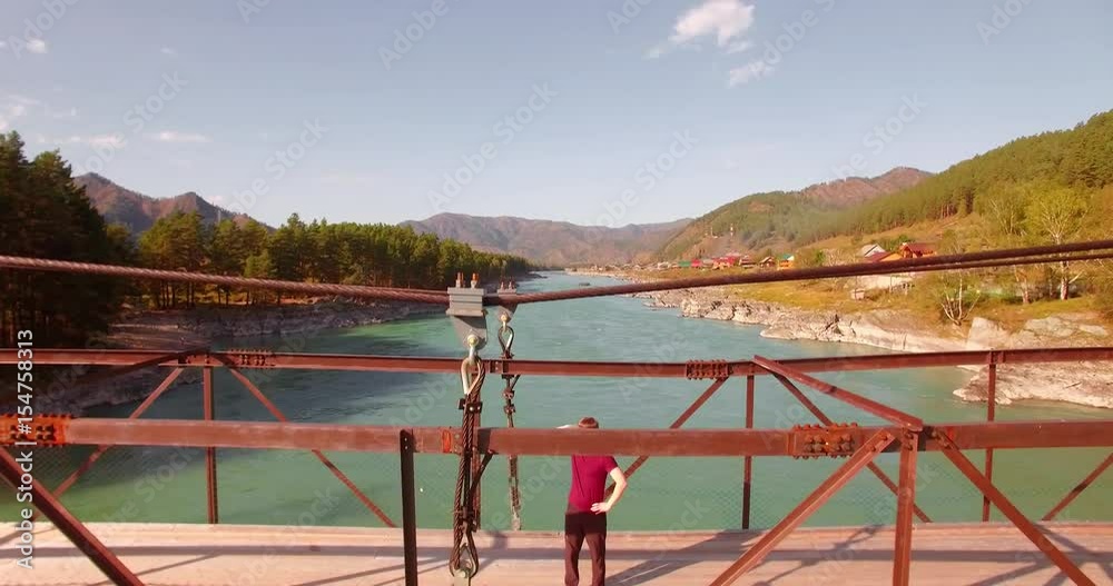 Aerial UHD view. Mid-air flight over young man tourist staying on a suspension bridge and watching. Fresh mountain river at sunny summer day. Vertical movement Aerial UHD view. Mid-air flight over young man tourist staying on a suspension bridge and watching. Fresh mountain river at sunny summer day. Vertical movement