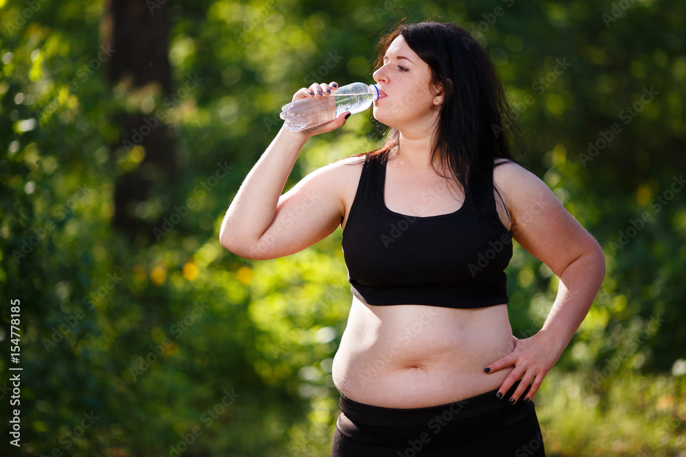 Young thirsty overweight woman drinking water after jogging in the park Young thirsty overweight woman drinking water after jogging in the park