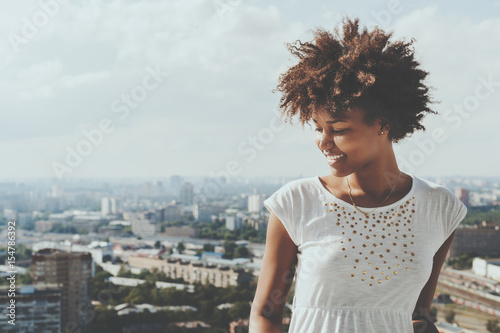 Cute laughing ebony girl looking down from balcony, smiling mixed teenage female with summer cityscape in background, young black lady in white dress with copy space zone for advertising or logo