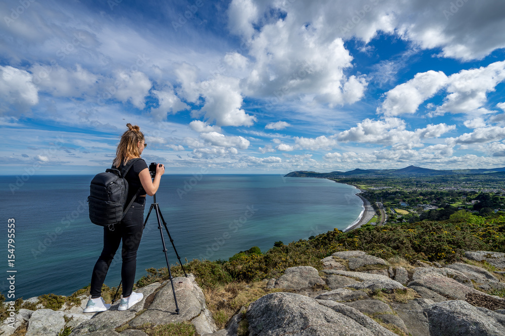 young beautiful girl female model photographer posing on top of the hill people embracing nature with open arms sea clouds blue sky tripod camera backpack sport clothes