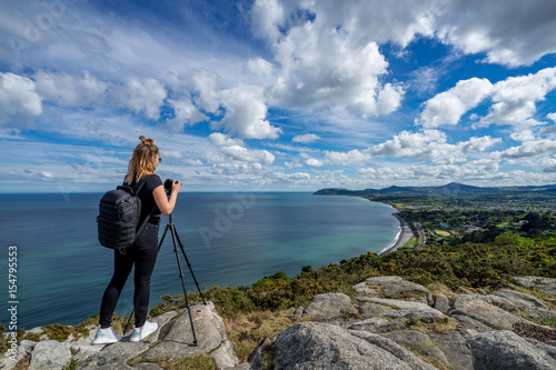 young beautiful girl female model photographer posing on top of the hill people embracing nature with open arms sea clouds blue sky tripod camera backpack sport clothes