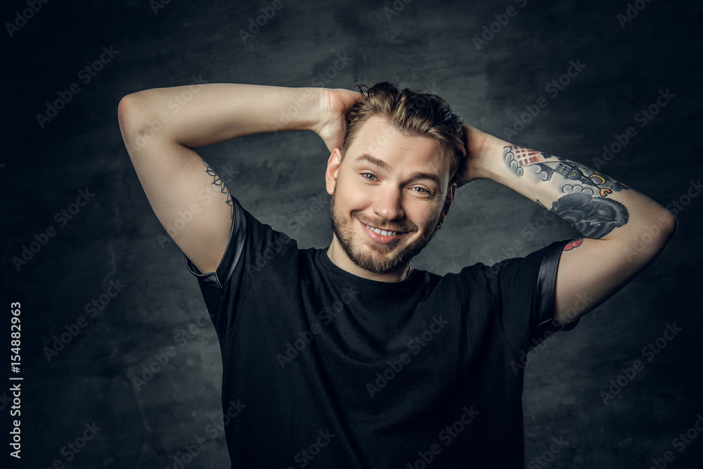 A positive hipster male dressed in a black t shirt holds head with tattooed arms.