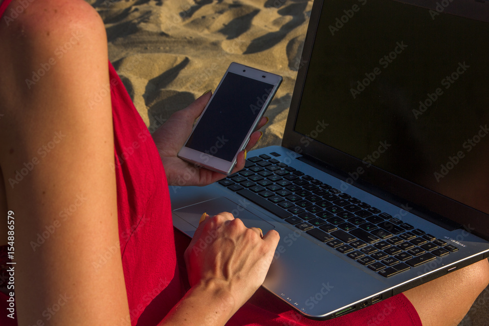 Naklejka premium Young woman in red dress with computer and smartphone on the beach. Freelance and downshifting concept.