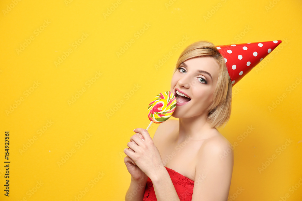 Beautiful woman in party cap holding lollipop on yellow background Beautiful woman in party cap holding lollipop on yellow background