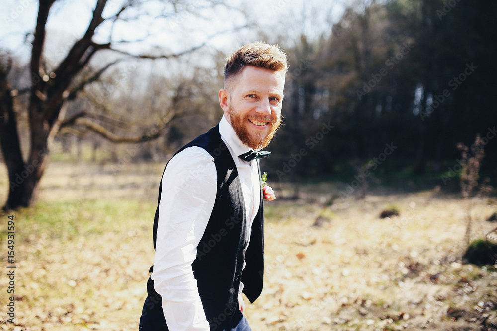 Handsome groom with red beard stands on the lawn full of sun Handsome groom with red beard stands on the lawn full of sun