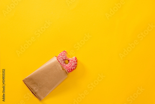 Top view of bitten doughnut in pink icing with paper bag isolated on yellow. Tasty donut background