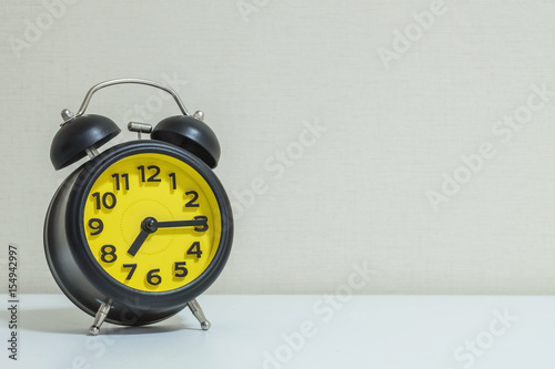 Closeup yellow and black alarm clock for decorate show a quarter past seven o'clock or 7:15 a.m.on white wood desk and cream wallpaper textured background tone with copy space
