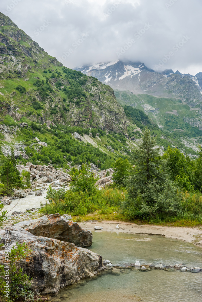 Summer cloudy landscape with Russian Caucasus snowy and woody mountains and small river at the foot of it..