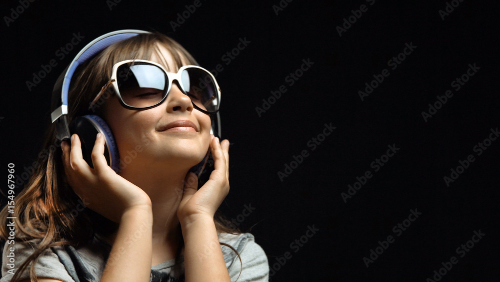 Studio shot of cute teenager girl wears headphones and sunglasses before black background, listening rhytmical music, youth freedom concept Studio shot of cute teenager girl wears headphones and sunglasses before black background, listening rhytmical music, youth freedom concept