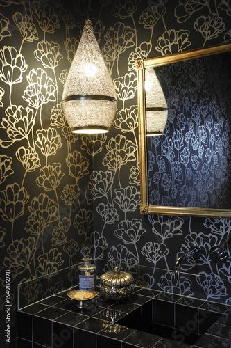 Black bathroom with black and gold floral wallpaper, Moroccan lamp with subdued light and black tiles photo