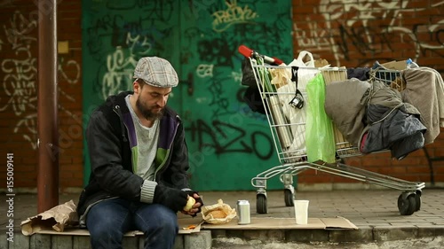 Homeless man sitting near his trolley with stuff and eating mandarin. photo