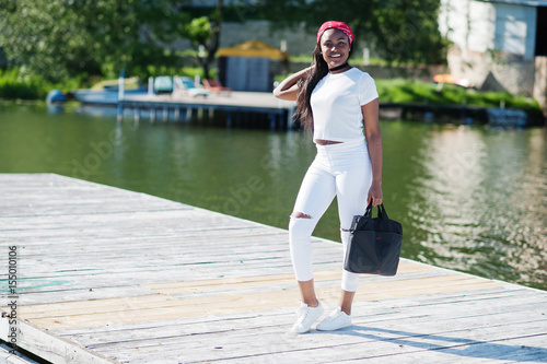 Portrait of stylish african american girl, wear on white clothes, with bag on hand against pier of lake. Street fashion of young black people. © AS Photo Family