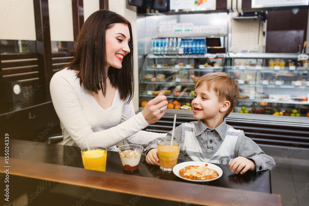 A young mother is feeding her little son with a spoon at a table in a cafe