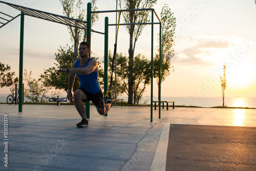 workout with suspension straps In the outdoor gym, strong man training early in morning on the park, sunrise or sunset in the sea background  © serejkakovalev