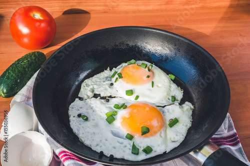 Fried eggs in pan, cucumber and tomato on wooden background