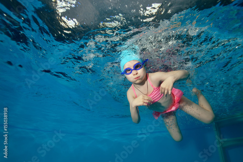 Little girl swims and plays sports underwater in the pool on a blue background and looks at me. Portrait. Close-up. Shooting under water. Horizontal orientation