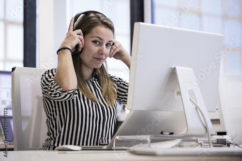 Giovane operatrice di call center con camicia a righe e cuffie è seduta alla sua scrivania nel suo ufficio guarda in macchina mentre si tiene le cuffie.