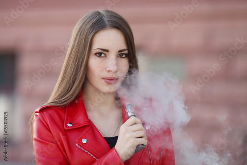 Woman in red jacket vaping hold modern e-cig device mouthpiece in lips.Quit smoking nicotine. Young ejuice smoker woman with electronic cigarette gadget, vaping device. photo